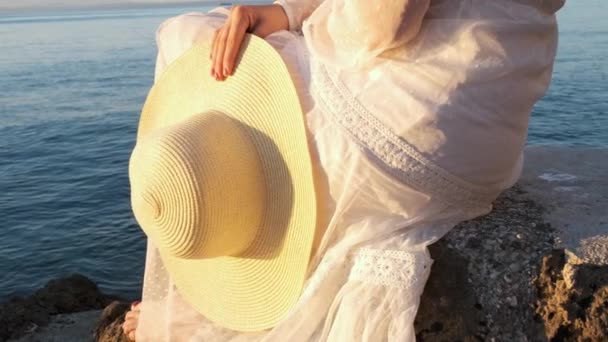 Mujer en el vestido blanco con sombrero en la mano sentada en la piedra cerca del mar al amanecer o al atardecer. — Vídeos de Stock