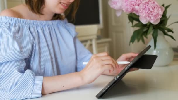 Junge, fröhliche Frau, die mit Bleistift am Tablet arbeitet, in modernem Interieur mit Pfingstrosen im Hintergrund. Freiberufler oder Designer, die zu Hause arbeiten. — Stockvideo