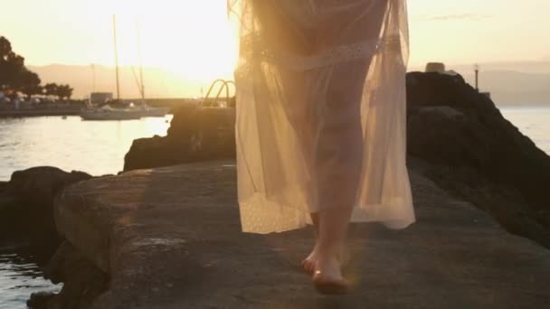 Una mujer camina a lo largo del muelle a lo largo del mar con un vestido blanco transparente al amanecer o al atardecer. — Vídeos de Stock