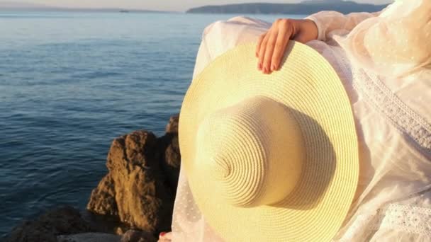 Frau im weißen Kleid mit Hut in der Hand sitzt auf einem Stein am Meer bei Sonnenaufgang oder Sonnenuntergang. — Stockvideo