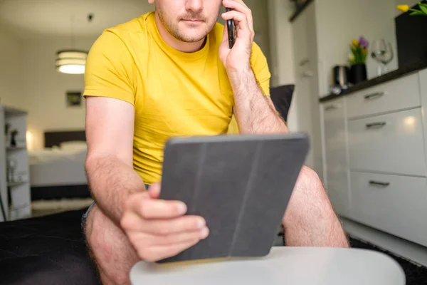 Человек в желтой футболке разговаривает через смартфон и использует планшет, сидя за журнальным столиком на современной кухне — стоковое фото