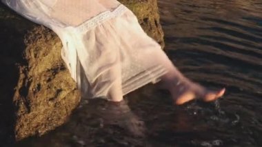 Beyaz elbiseli bir kadın gün doğumunda bacakları sarkan bir su ile. Gün batımında kayadan gelen ayaklarla rahatlar ve denize dokunurlar.. 