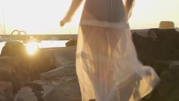 Egy nő fehér ruhában és fekete testben szalad a mólón a tengerhez naplementekor vagy naplementekor. Élvezze a gyönyörű kilátást és a kikapcsolódást — Stock videók