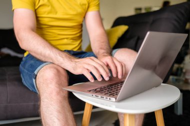Modern oturma odasında oturan sarı tişörtlü adam dizüstü bilgisayarda yazıyor. Evde çalışan serbest çalışan biri.