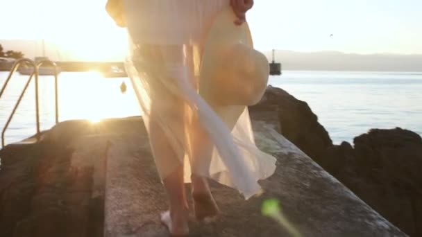 Жінка в прозорому одязі з капелюхом в руці йде вздовж пірсу біля моря на сході або заході сонця — стокове відео