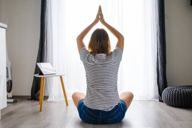 Genç bir kadın oturma odasında meditasyon yapıyor ya da yoga yapıyor. İnternetteki eğitim uygulaması tableti kullanıyor. 