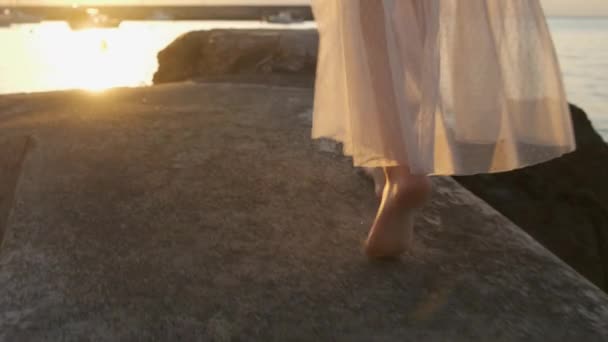 Жінка в білій сукні, що рухається вздовж кам'яного узбережжя біля моря на сонячному світлі — стокове відео