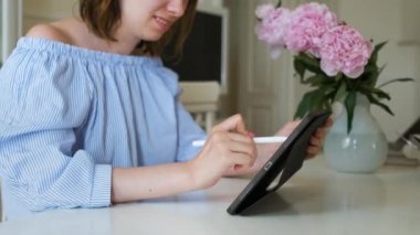 Tablet üzerinde çalışan neşeli genç bir kadın modern iç kısımlarda kurşun kalemle arka planda şakayıklar var. Evde çalışan serbest ya da tasarımcı. 