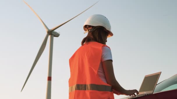 Um engenheiro de moinho de vento supervisiona o trabalho das turbinas eólicas no laptop que está perto do carro. Produção de electricidade ecológica. — Vídeo de Stock