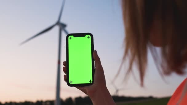 Női mérnök kezében egy okostelefon chroma kulcs a háttérben a szélturbina naplementekor. — Stock videók