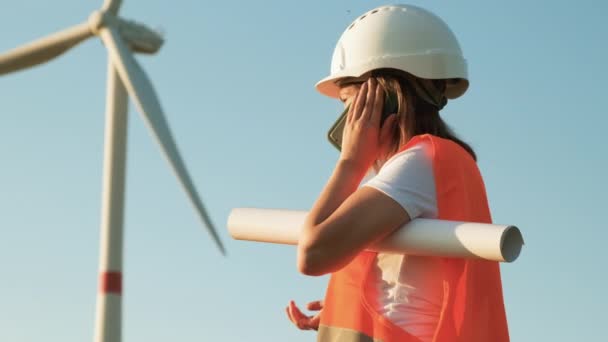 Ingenjör i hårdhatt och skyddsväst har ett planprojekt för uppförande av vindkraftverk i fält och kallar mot bakgrund av väderkvarnar. Elproduktion. Alternativ — Stockvideo