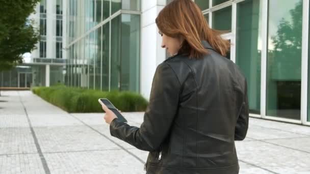 女性はビジネスセンターの近くに立って、クライアントとのビデオ通話のためのスマートフォンを使用します — ストック動画