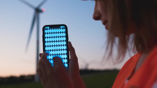 Női mérnök kezében egy okostelefon krómkulccsal, és hogy zoom gesztus a háttérben a szélturbina naplementekor. — Stock videók