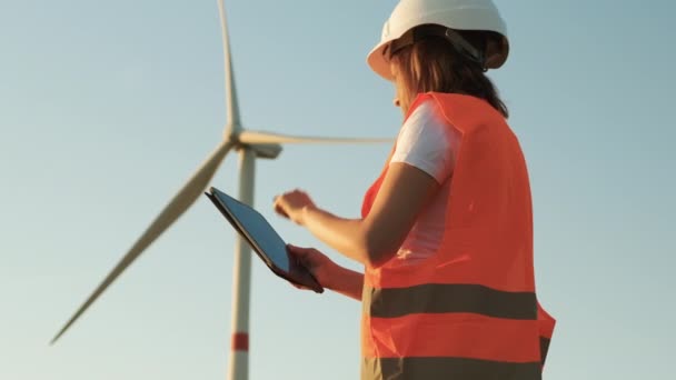 Kobieta inżynier w kasku i pomarańczowej kamizelce kontroluje działanie turbiny wiatrowej za pomocą tabletu na tle nieba — Wideo stockowe