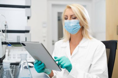 Blonde woman in protective lab mask and gloves writing down chemical experiment outcomes on a tablet in a modern laboratory with HPLC, flasks, and other instruments