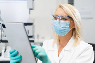 Middle aged female scientist wearing mask and lab coat using tablet in laboratory for research and analysis 