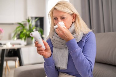 Middle aged blonde woman lying on couch at home, blowing her nose into a tissue and checking her temperature with a thermometer. Concept of flu, cold, fever, and home recovery.