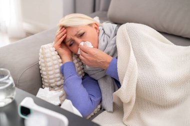 Burun tıkanıklığı olan, ilaç ve termometreyle çevrili, kanepede dinlenen hasta sarışın kadın. Mevsimsel hastalık kavramı, soğuk, evde dinlenme ve iyileşme..