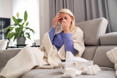 Evdeki koltukta oturan orta yaşlı sarışın kadın üşümüş ve başı ağrıyormuş. Battaniyeye sarılmış, iyileşirken dinleniyor..