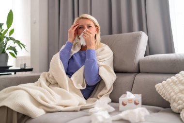 Orta yaşlı, sarışın bir kadın evinde kanepede oturuyor. Baş ağrısı ve nezle yüzünden başını tutuyor..