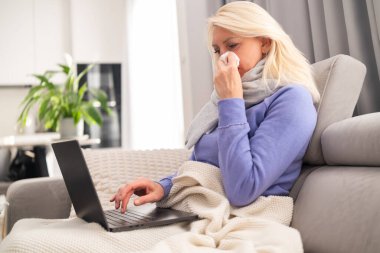 Blonde middle aged woman sitting on a sofa in a cozy room feeling unwell and watching a film on her laptop while staying at home