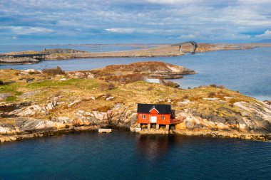 Norveç kıyılarının geniş fiyortlu suları arasında kayalık kahverengi bir adaya kurulmuş küçük bir ev. Dağlar Norveç 'in dramatik doğal güzelliğini vurgular 