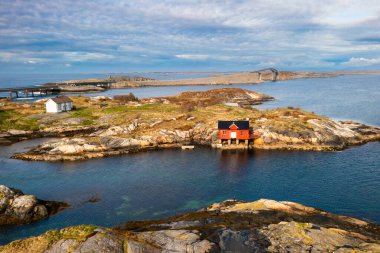Norveç kıyıları boyunca uzanan geniş fiyortlu sularla çevrili kayalık kahverengi bir adaya tünemiş küçük ahşap ev, Norveç 'in dramatik doğal güzelliğini vurgulayan yüksek dağlarla çevrelenmiş..