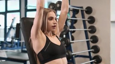 Bayan, kollarını dirseğindeki dambıllarla dikey olarak eğiyor. Sarışın kadın pazılarını güçlendiriyor ve hareketlerini sergiliyor. Tutuşunu güçlendiriyor.