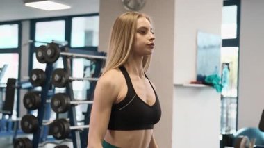 Kadın geniş spor salonunda ayakta duran halterleri kaldırıyor. Sarışın atlet deltoidler ve üst göğüs kasları arasında duruyor.