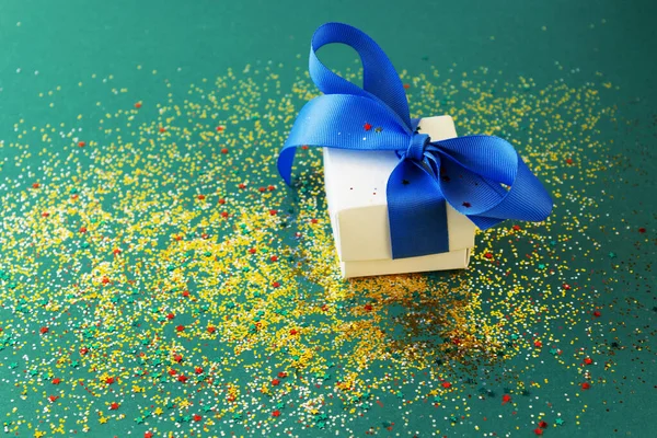 Yeşil arka planda mavi kurdeleli beyaz hediye kutusu. Şenlikli konfeti yeni yıl havası yaratır..