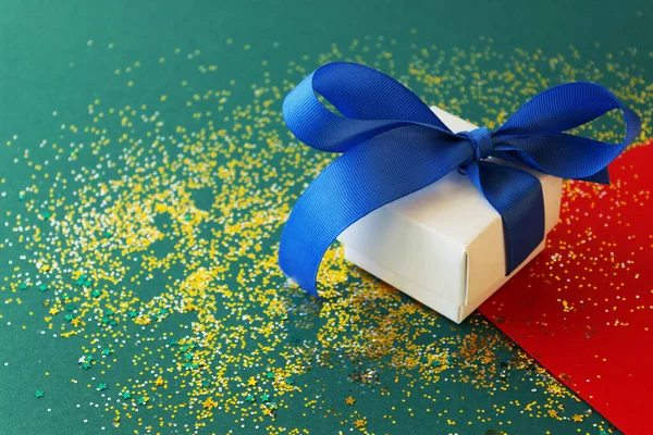 Yeşil arka planda mavi kurdeleli beyaz hediye kutusu. Şenlikli konfeti yeni yıl havası yaratır..