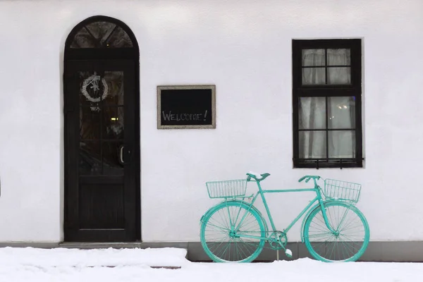 Duvarın yanında mavi bir klasik bisiklet duruyor. Kapılar Noel arifesinde süslenir. Duvarında selam yazan bir pano. Minimalizm.