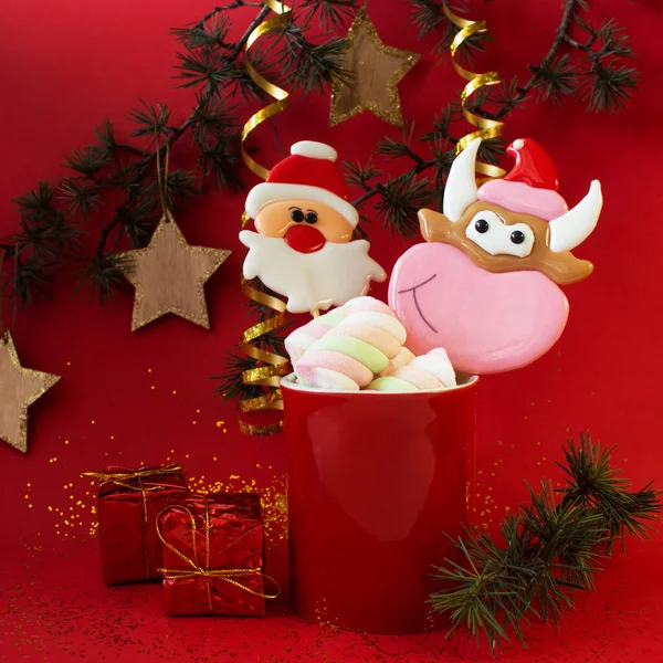 Noel şekerlemeleri ve marşmelovlar kırmızı bir bardakta, yeni yıl oyuncakları ve hediye kutularıyla süslenmiş ladin dalının arka planında..