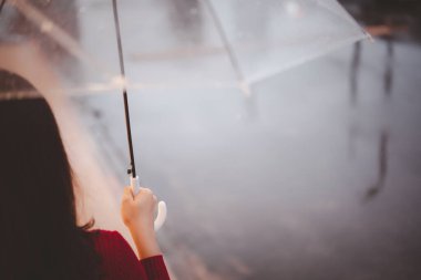 Yağmur yağarken ve elinde şemsiyeyle yol kenarında duran kırmızı kazaklı güzel bir Asyalı kadın. Yağmurlu saatlerde şehir merkezinde gezen neşeli Asyalı kadın portresi yağmurda.