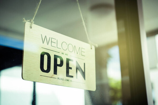 Vintage retro wooden sign with words "Welcome We Are Open, Please Come In" hanging in front of the restaurant glass door close up with copyspace. Welcome sign on the store window close up.