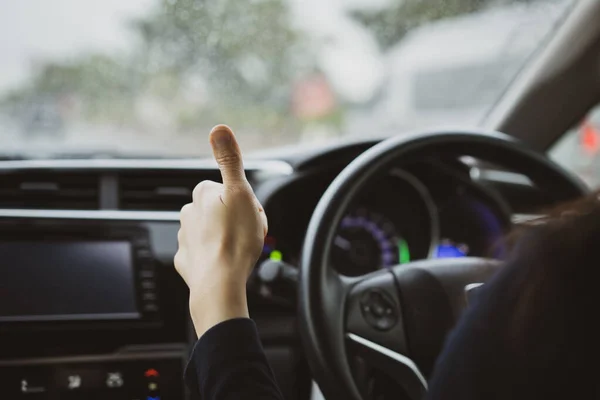 Unrecognizable Asian woman driver showing her thumb up sign during driving a car in the rain in the meaning of safety and responsibility driving concept.