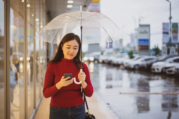 Happy Asian woman in a relax smart casual dressing using a smartphone while walking the in rain.