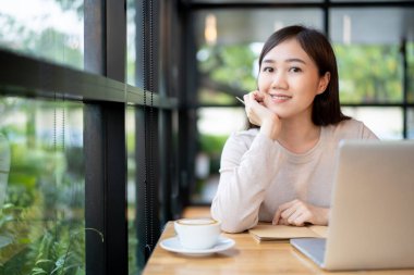 Çalışan Asyalı güzel bir kadın kahvecide ya da kafede içki içiyor. Bilgisayarına ya da bilgisayarına yazı yazan neşeli bir internet blogcusu ve yakın mesafeden kahve içiyor. Modern yaşam tarzı.