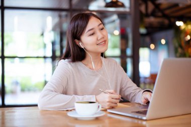 Çalışan Asyalı güzel bir kadın kahvecide ya da kafede içki içiyor. Bilgisayarına ya da bilgisayarına yazı yazan neşeli bir internet blogcusu ve yakın mesafeden kahve içiyor. Modern yaşam tarzı.