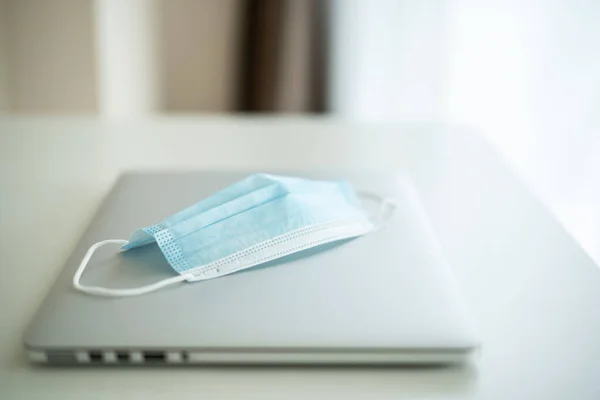 Hygiene protective face mask and laptop computer on the table in home office close up with copyspace. Healthcare and new normal concept in COVID-19 or Coronavirus 2019 pandemic.