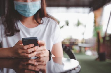 Kafe ve restoranda dinlenirken yüz maskesi takan bir kadın akıllı telefondan fotokopi çeken bir kadın. Asyalı genç yetişkin dışarıda kalırken koruyucu yüz maskesi kullanıyor..