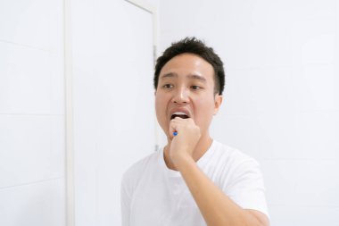 Beyaz gömlekli Asyalı genç bir adam sabah yataktan kalktıktan sonra aynanın karşısında dişini fırçalıyor, ağız sağlığı ve sağlık hizmetleri. Diş sağlığı konsepti.