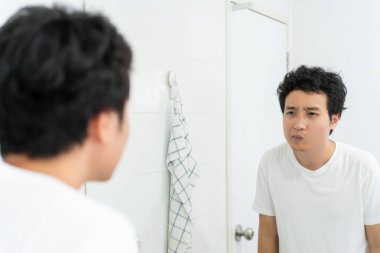 Beyaz tişörtlü mutlu Asyalı genç adam sabahları tuvalette aynada kendine bakıyor. İşe gitmeden önce yüzünü ve vücudunu hazırlayan bir adam. Günlük yaşamda rutin aktivite.