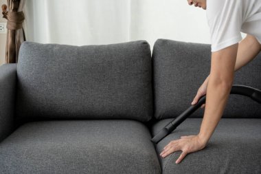 Tanımlanamayan Asyalı bir temizlikçi vakum makinesi kullanıyor oturma odasındaki koltuğun tozunu temizliyor. Temizlik ve temizlik için evini temizleyen bir adam. Elektrik süpürgesi ve ev işi.