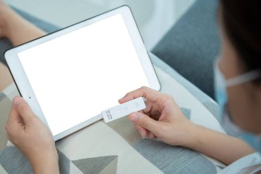 Boş ekran kırpma yolu ile izole edilmiş modern tablet. COVID-19 antijen testi sonucu pozitif çıktıktan sonra bir doktorla çevrimiçi video görüşmesi için tablet kullanan Asyalı genç kadın.