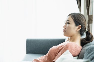Oturma odasındaki kanepede dinlenen mutlu Asyalı genç kadın. Kaygısız Asyalı kadın bir ev işi yaptıktan sonra dinlenmek için kanepeye oturuyor..