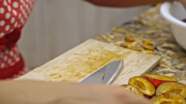 Kadın taze erikleri ikiye bölüyor ve mutfaktaki çukurları temizliyor. Yakından bakıldığında meyve hazırlama sürecinde ellere odaklanmış.