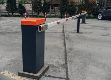 Turuncu kapaklı ve siyah direkli bir park engeli, beton bir alana kurulmuş. Kapı kısmen açık, bu da park alanına erişimi gösteriyor..