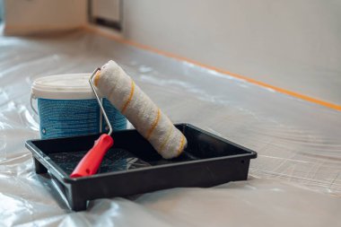 Bir boya silindiri ve tepsi, bir kova boyanın yanında, bir ev yenileme projesi için hazırlandı. Zemin koruyucu plastikle kaplı..