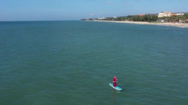 Sörf üzerinde hediye paketi olan Noel Baba. Noel Baba denizde sörf tahtasında. Adam Noel Baba kostümüyle spor yapıyor. Noel ve Yeni Yıl konsepti. Noel Baba 'ya mavi ve yeşil okyanusta kürek ve hediye çantasıyla havadan izleyeceğiz.